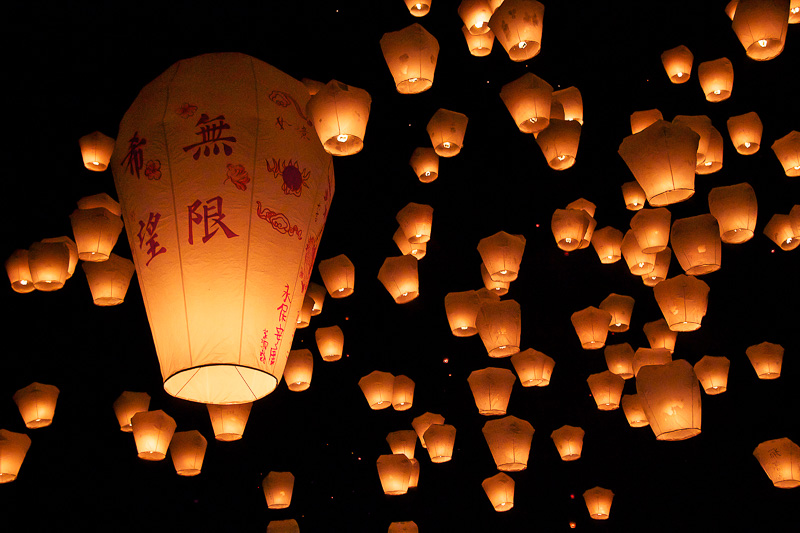 Many lanterns going up into a black sky with one especially big one and each one has a small fire inside