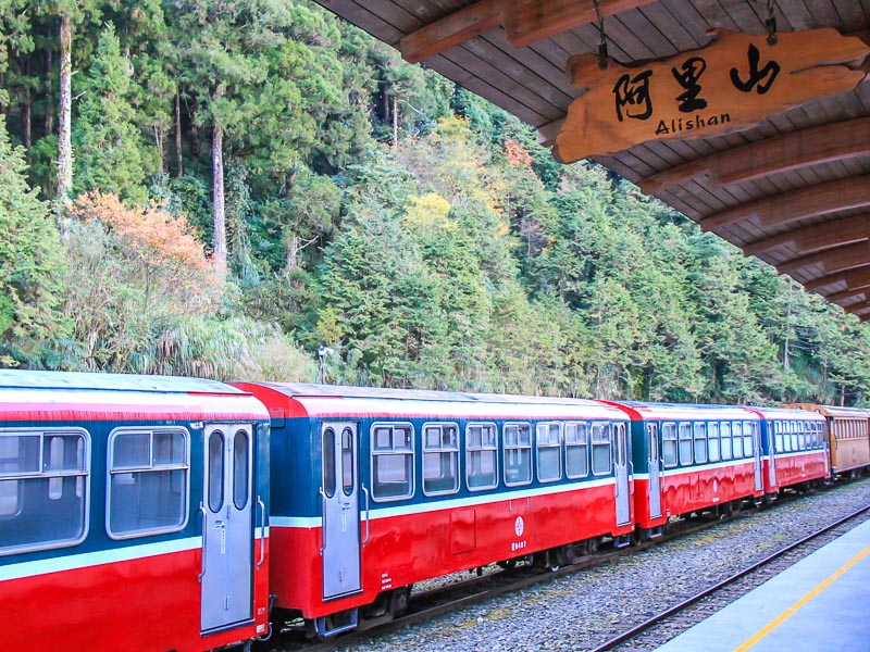 A red train parked in a station with trees behind, and a sign that says Alishan