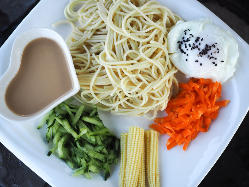 Looking straight down at a white plate with noodles, sesame sauce, vegetables, and egg