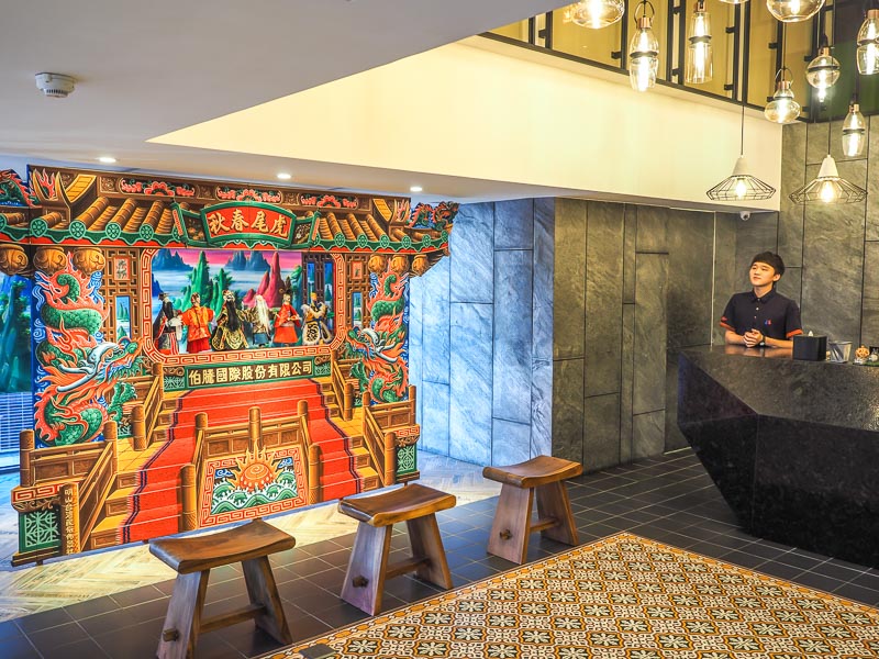 A hotel lobby with a clerk at the desk on the right, traditional Taiwanese hand puppet stage on the left, and hanging lights above