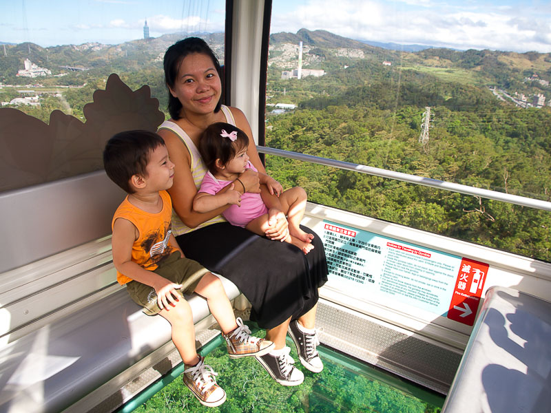 A Taiwanese mother and her two young kids sitting inside a gondola with glass floor and sides and view of Taipei 101 in distance 