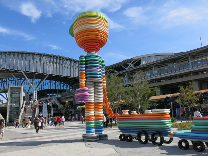A plaza in front of a large train station with a colorful statue