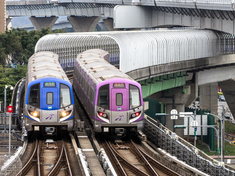 Two Taoyuan Airport MRTs passing each other on tracks before a tunnel, one is blue and one is purple