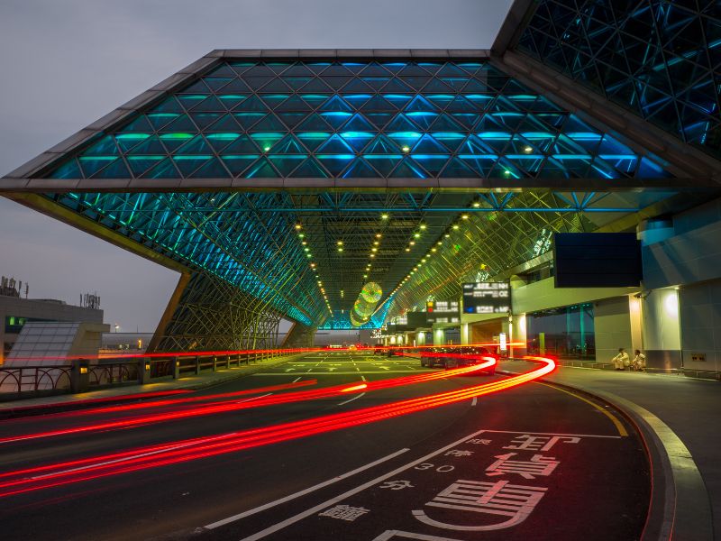 Glass roofed entrance to Taoyuan Airport at night, with flowing red lights of a car driving up the entrance
