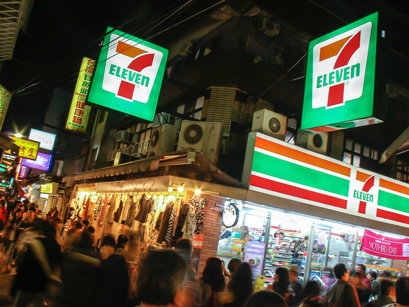 A 7-Eleven at night with many people walking on the dark street outside it