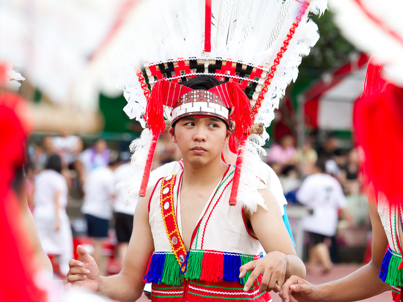 A young Taiwanese aboriginal man wearing traditional clothing and large decorated hat holding hands with others while doing a dance performance