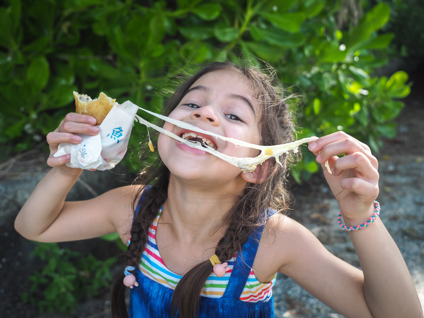 A young girl holding up a fried cheese roll in a paper bag, with long melted cheese strings from her other hand to her mouth