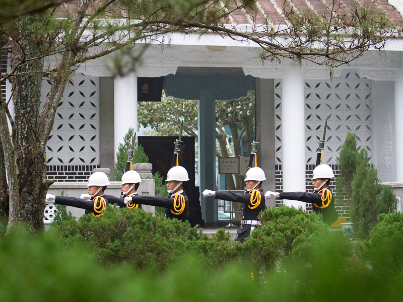 Soldiers marching with guns in front of Cihu Mausoleum, framed by tree branches at the top and bushes at the bottom