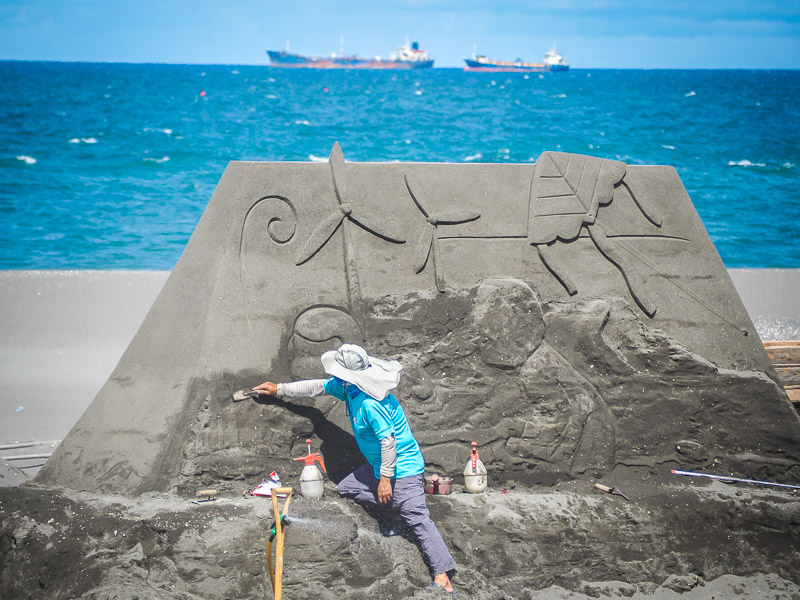 A man sculpting a large sandcastle with black sand and ocean behind