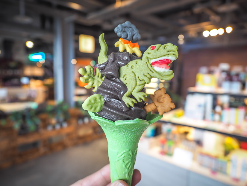 A hand holding up a green ice cream cone with black soft serve ice cream and a dinosaur shaped cookie