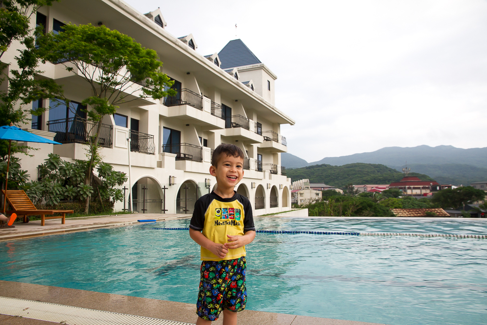 A young boy in bathing suit and swimming shirt beside a pool with large white hotel behind it