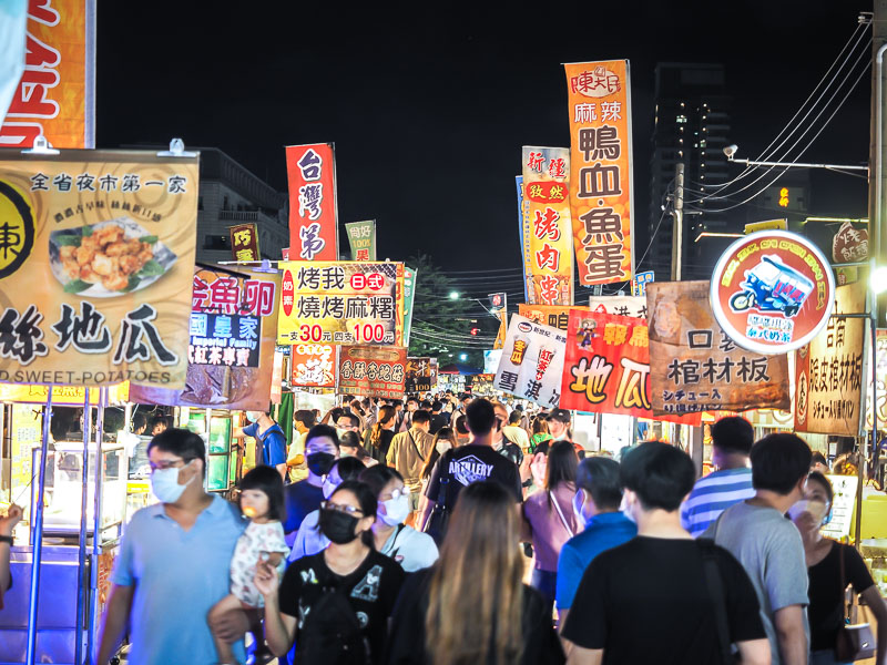 A crowded aisle between food vendors in Garden Night Market with food stall signs sticking up into the air