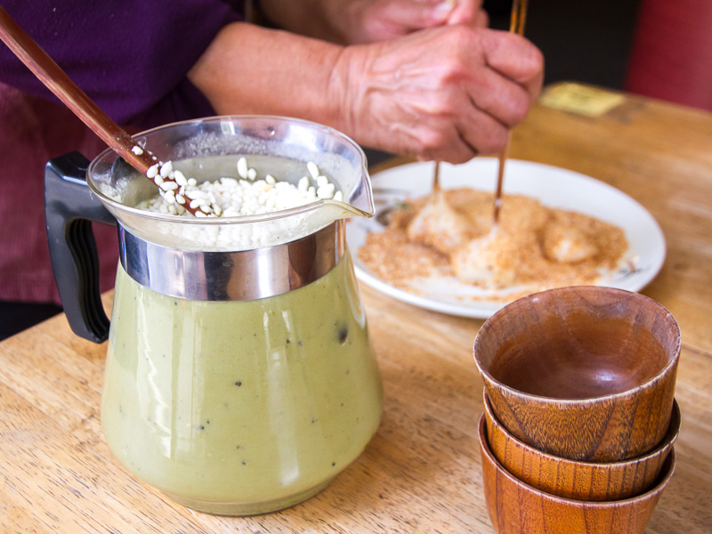 Two hands of an elderly person using chopsticks to dip some mochi balls in peanut powder and a pitcher of Hakka pouned tea with rice puffs on the top