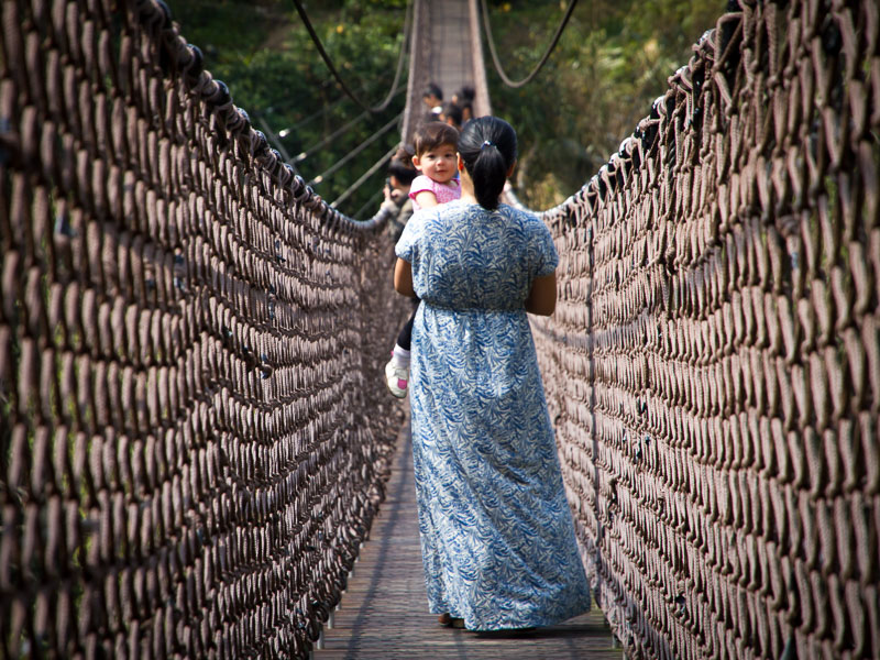A woman's back crossing a suspension bridge, holding a young girl who is facing the camera