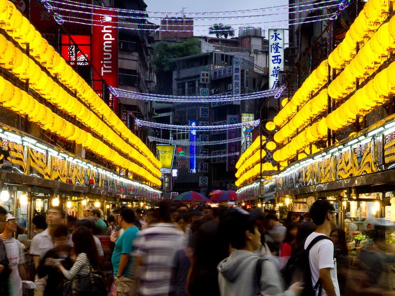 Crowds of people blurred inside Keelung Night Market at night, with rows of yellow lanterns above
