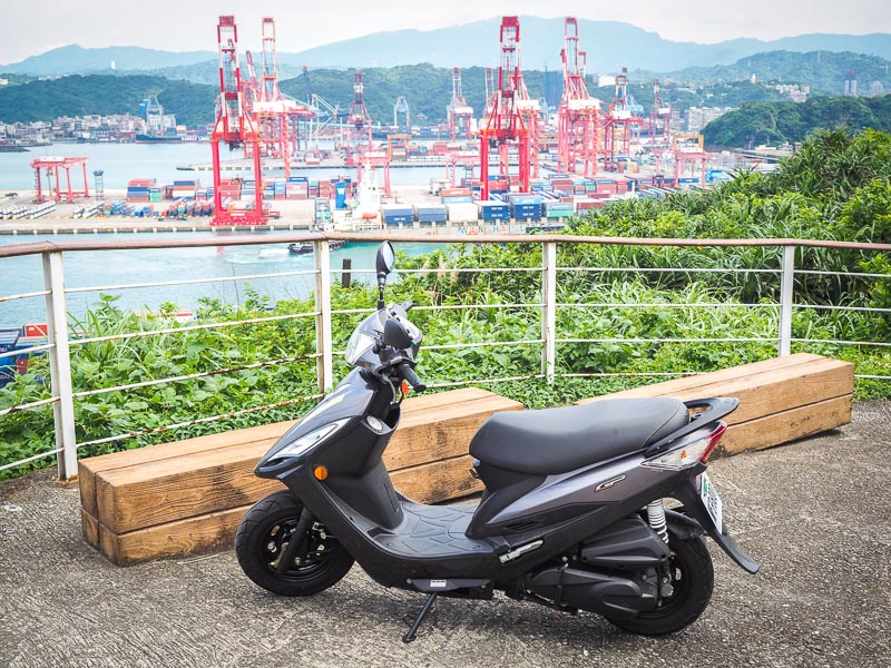 A scooter parked with a view of some port cranes in the background