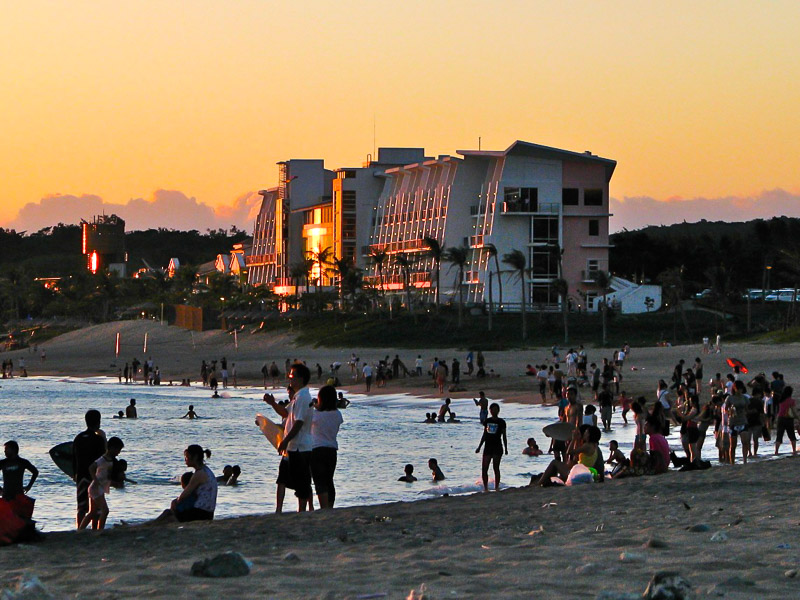 Crowds of people on a beach at sunset time, with a large resort on the beach reflecting the sunlight