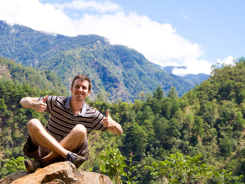 A young man sitting crosslegged on a rock giving two thumbs up with beautiful mountain scenery behind him
