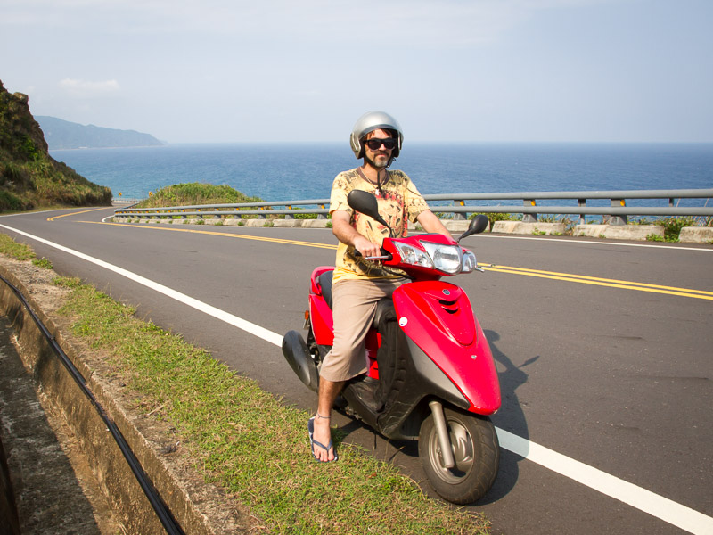 Nick Kembel on a red scooter on the highway with the coast of Kenting visible behind him