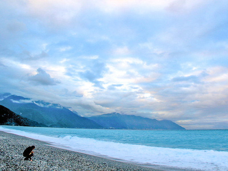 A person wearing all black and kneeling down on a gray pebble beach with coastal mountains in the background
