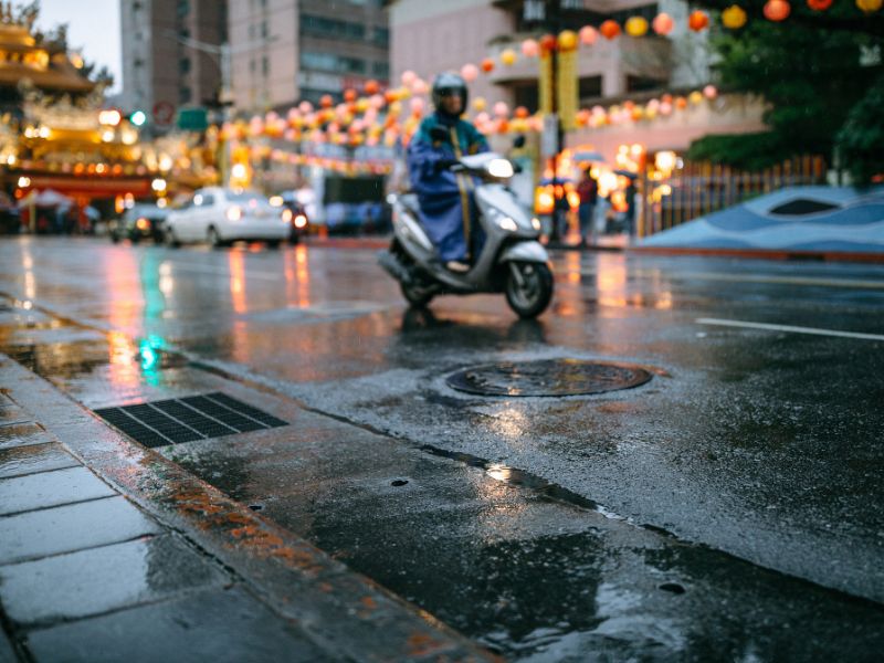 A scooter driving on a wet street in the rain, with temple lanterns in the background