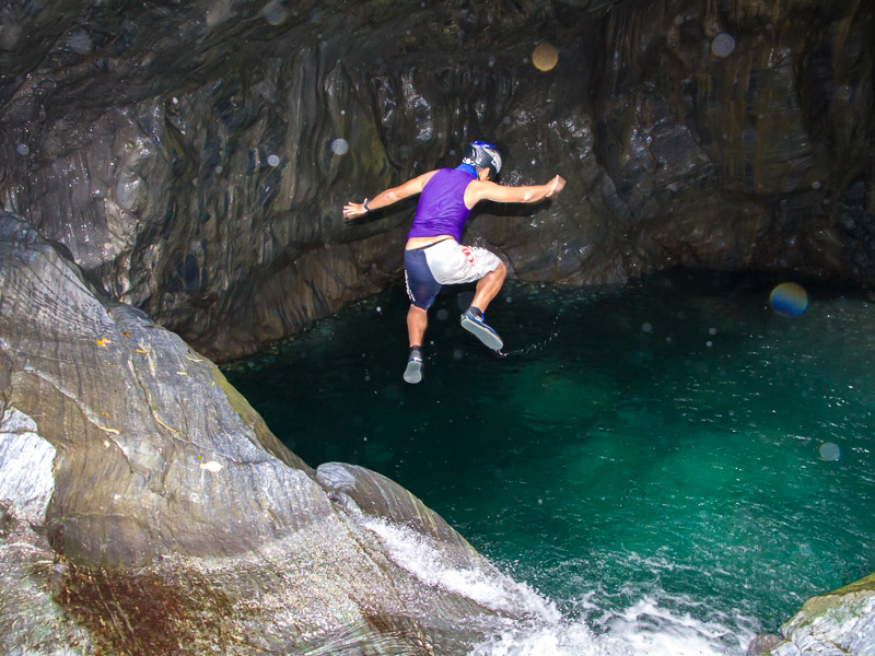 A man wearing helmet, purple tank top, swimming suit, and river tracing shoes jumping off the top of a waterfall into a green pool of water surrounded by cliffs