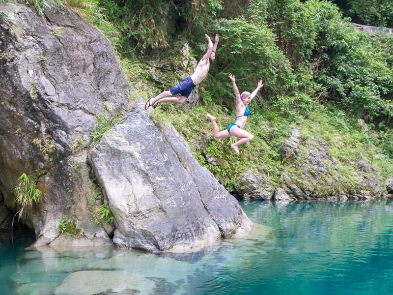 A man and woman jumping off a small cliff into clear green-blue water