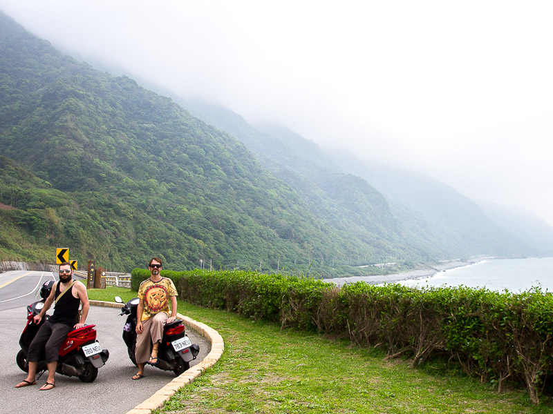 Nick Kembel and his friend standing beside scooters parked at the side of Highway 11 in Hualien with coastal mountains behind them