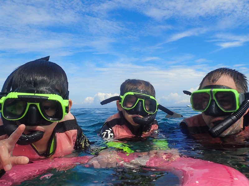 Nick Kembel and his two kids wear snorkeling hear and facing the camera while floating in the sea and holding a red floaty