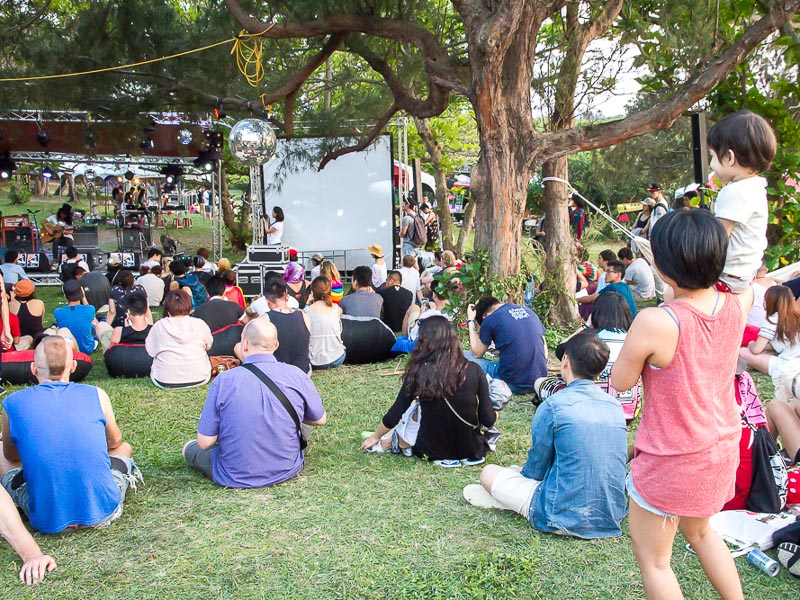 A small crowds of people sitting in the grass watching a concert, with a Taiwanese woman holding a toddler up to see