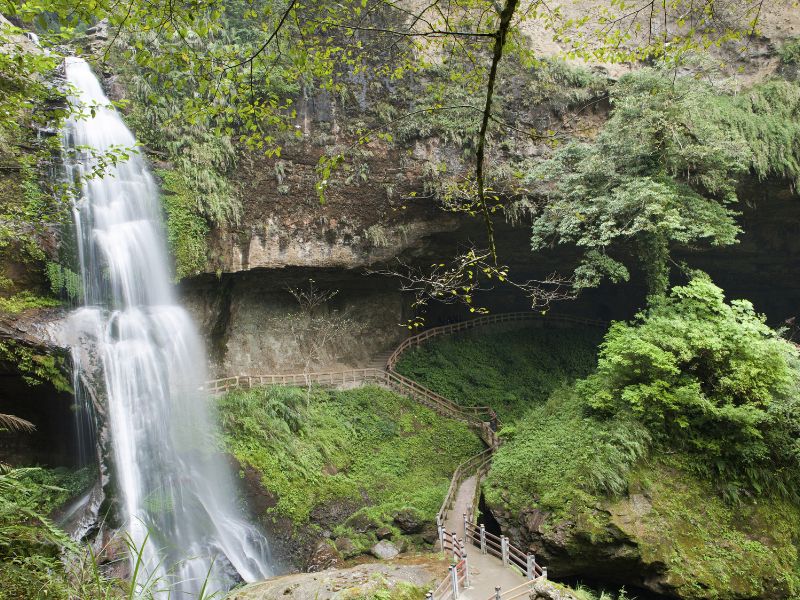 A waterfall pouring off the side of a cliff surrounded by forest and walking trails below