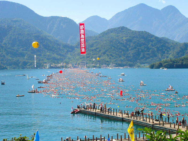 Thousands of people swimming across a lake, with a dock leading out onto the lake and balloons in the sky