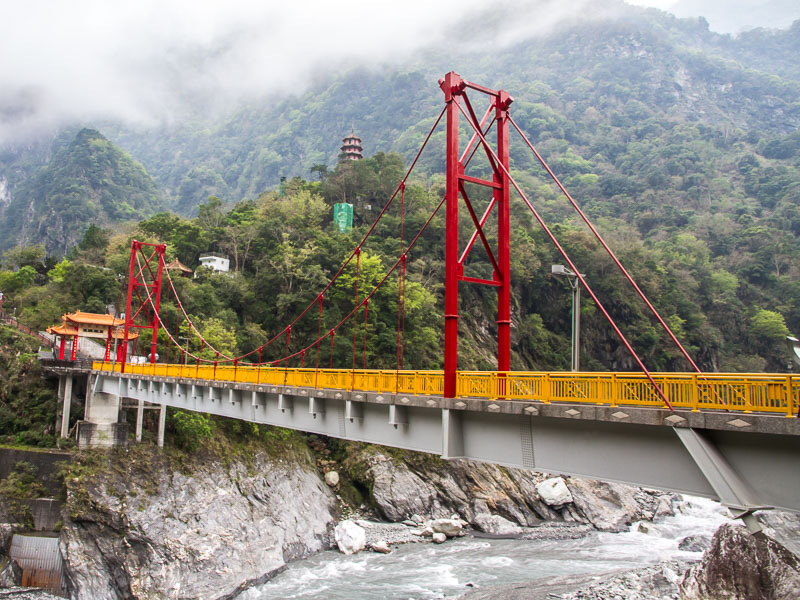 A red, orange, and cement bridge across a river in a beautiful valley with misty clouds above