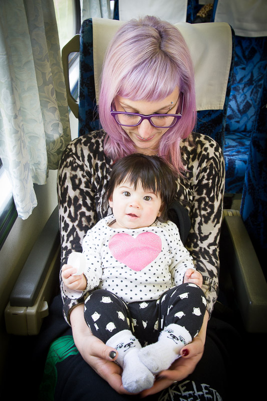 A young woman with pinkish purple hair holding a baby girl with a heart on her shirt while sitting on a train in Taiwan