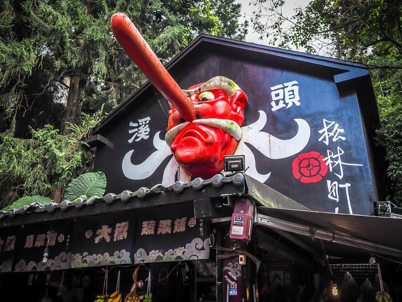 A Japanese looking wooden building with a red monster face statue with a long nose on the side