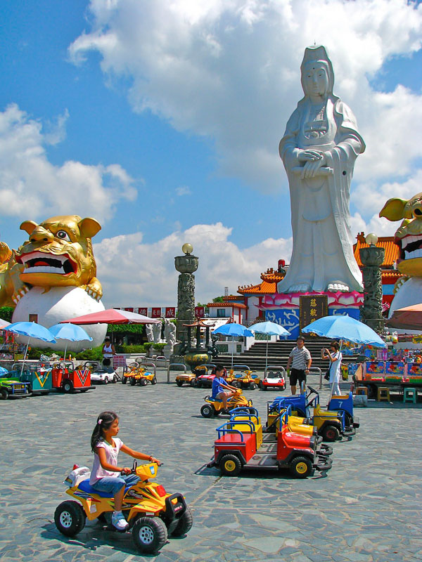 A tall white Kuanyin statue with some kids riding little cars around in the foreground