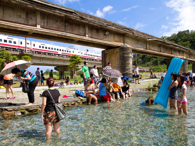 Many people standing or sitting beside a clear water cold spring, with a train passing by on a bridge above them