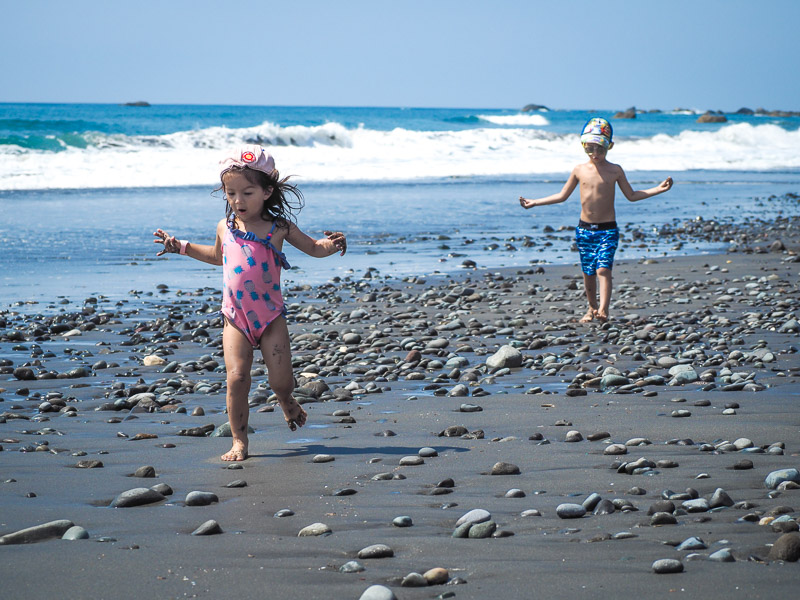 Two kids running down a gray sand beach beside the ocean