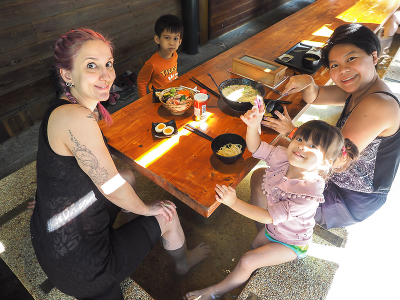 A family sitting a table eating noodles with their feet soaking in hot spring water below