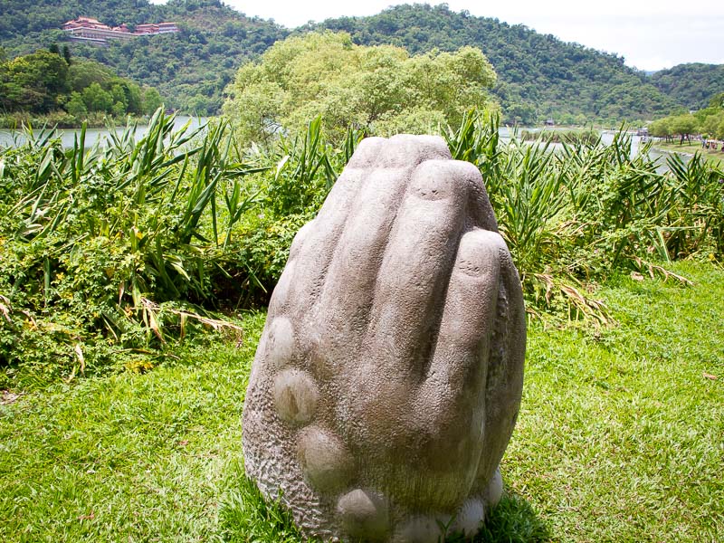 A statue of two praying hands surrounded by greenery and a lake in the background