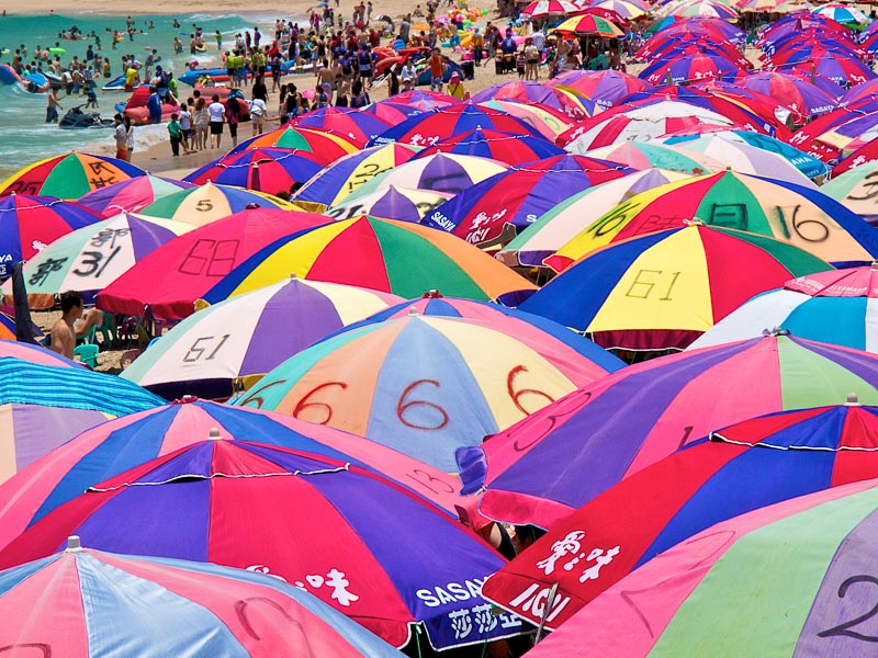 A huge collection of beach umbrellas packed tightly together with some swimmers on the beach in the background