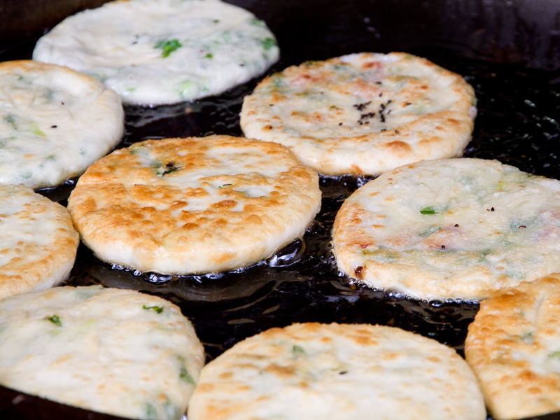 Several round green onion cakes frying in oil on a large black grill