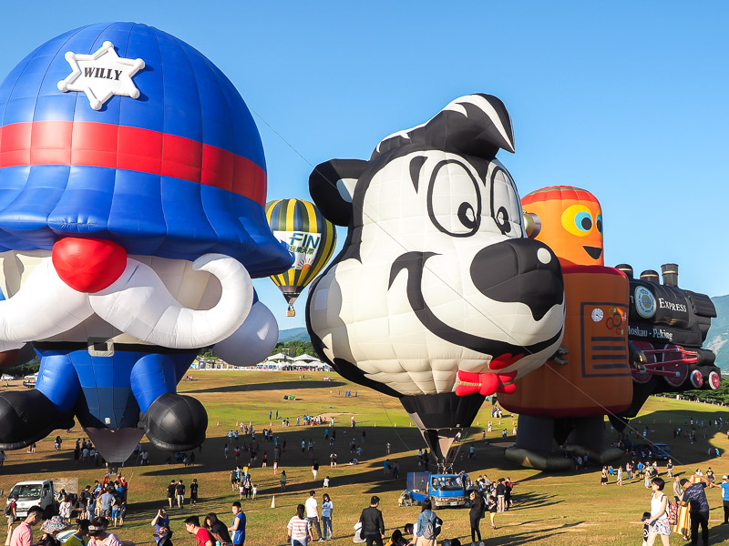 A row of cartoon shaped hot air balloons with people sitting on a hill looking at them